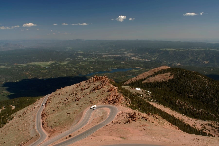 Photo overlooking Pikes Peak Highway in Colorado, including a few vehicles, red dirt, green trees, and water in the distance. 