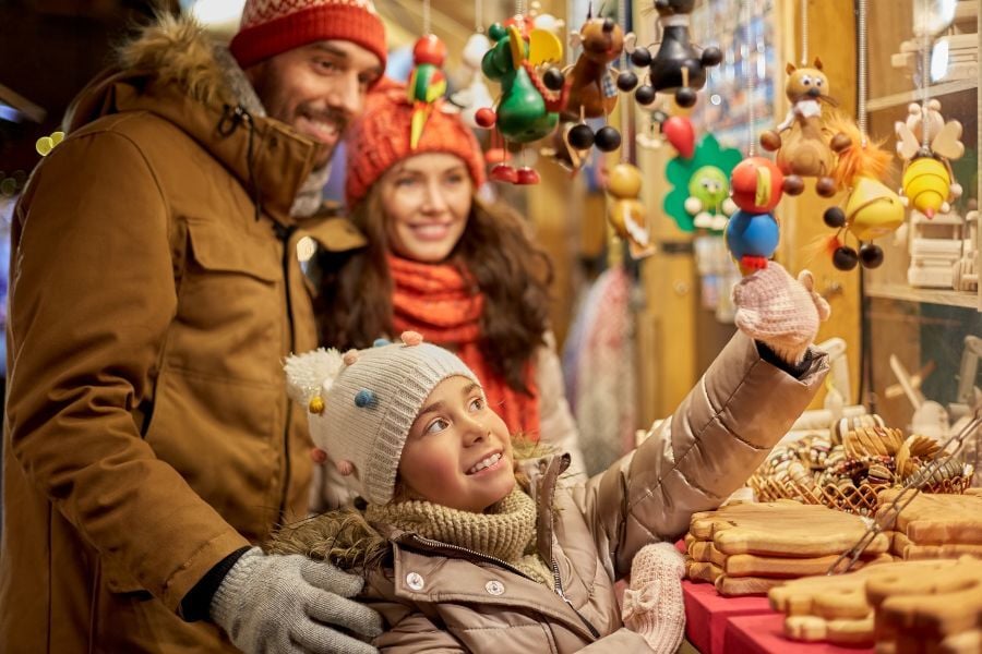 Girl in polka dot beanie hat admires ornaments at Christmas market with smiling mom and dad.
