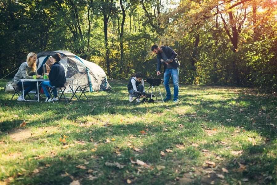 Family of four camping out in green clearing in forest with tent, table, and charcoal grill. 