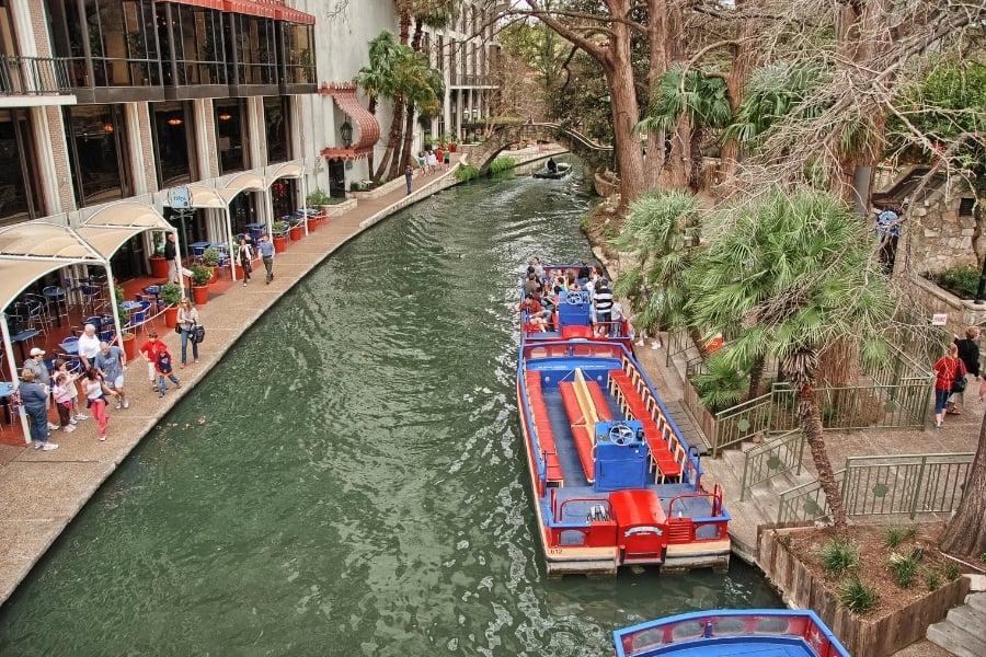 View of red and white boat floating near trees at the San Antonio river walk with people walking around.