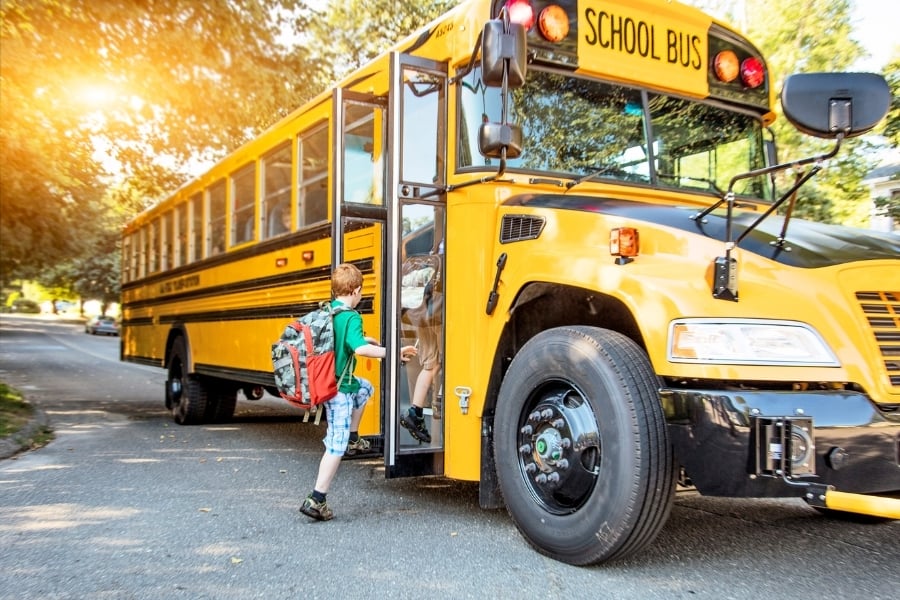 Red-haired boy in green shirt and blue plaid shorts wearing backpack steps onto yellow school bus on residential street with sun flare shining through the trees.