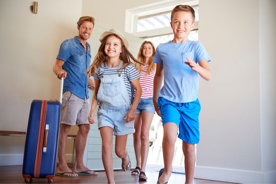 young couple with little boy and girl in blue clothing running happily into vacation rental with suitcases