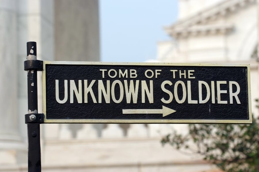 Photo of black and gold sign in front of historic buildings in D.C. that reads Tomb of the Unknown Soldier with arrow pointing to the right. 