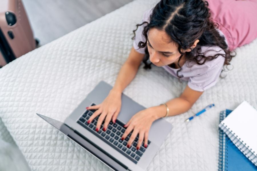 Woman wearing pink and purple lays on white bed typing on laptop. 