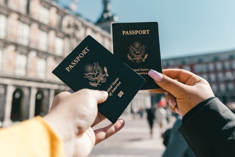 Two people holding U.S. passports up together in front of large European buildings in the background. 