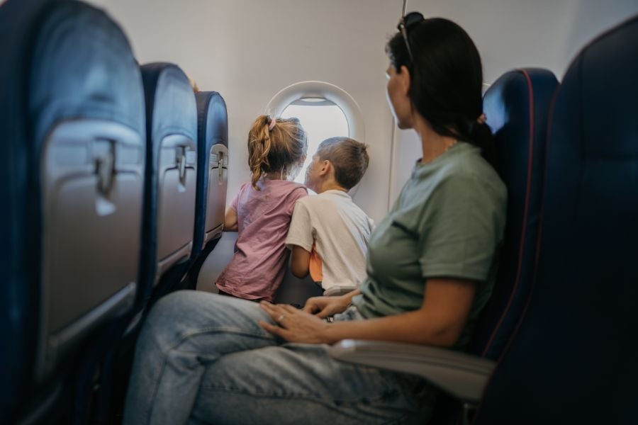 Mom in green t shirt sits with a young boy and girl on airplane looking out window.