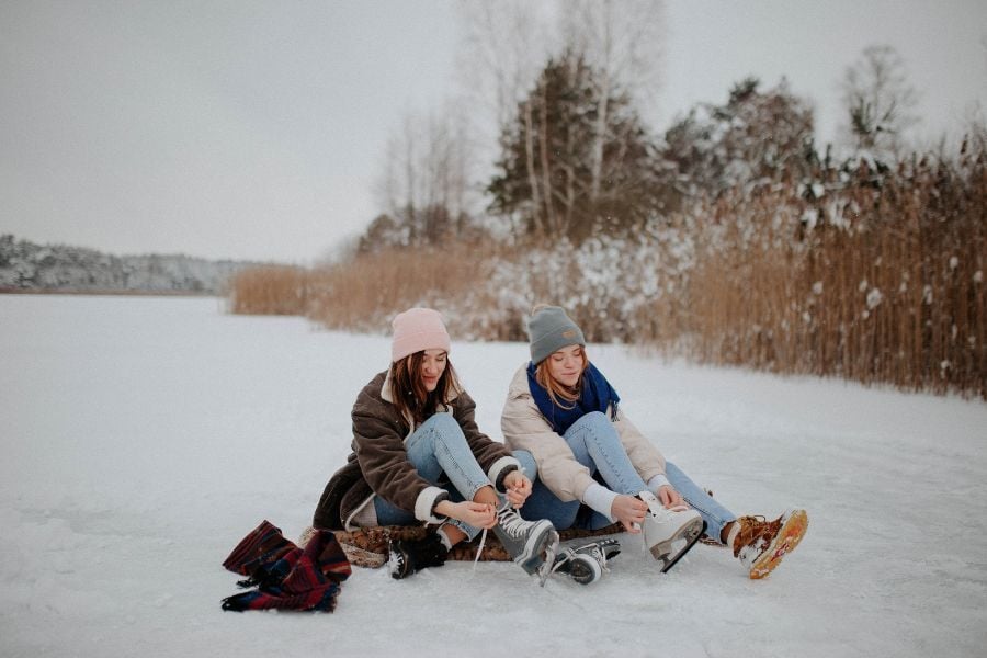 Two women in winter beanies and coats sit on icy pond and tie up skates.