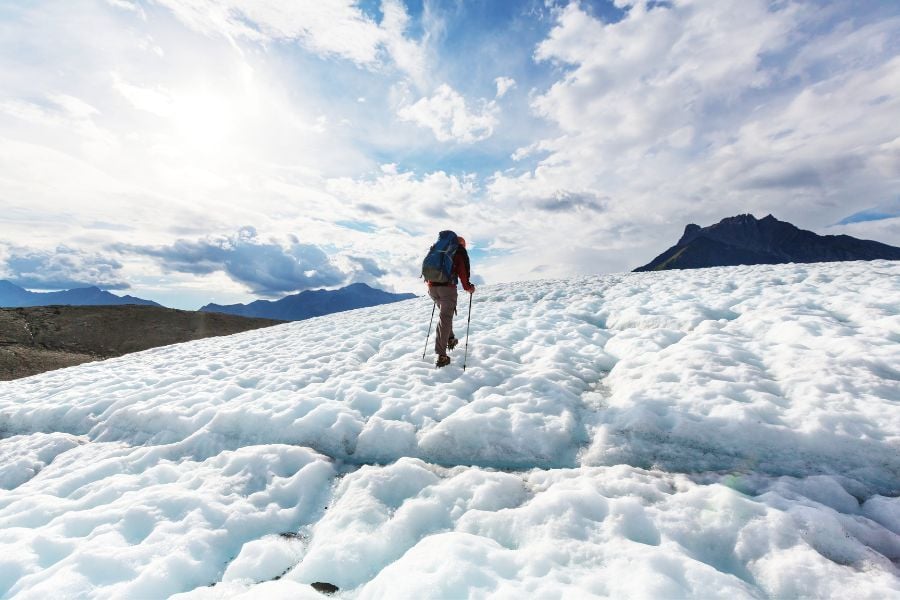 Man in red wearing blue hiking pack hikes up icy mountain using walking poles