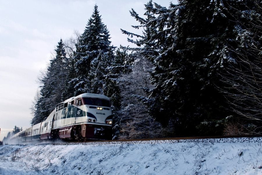 Amtrak train steaming on track through snow and evergreens.