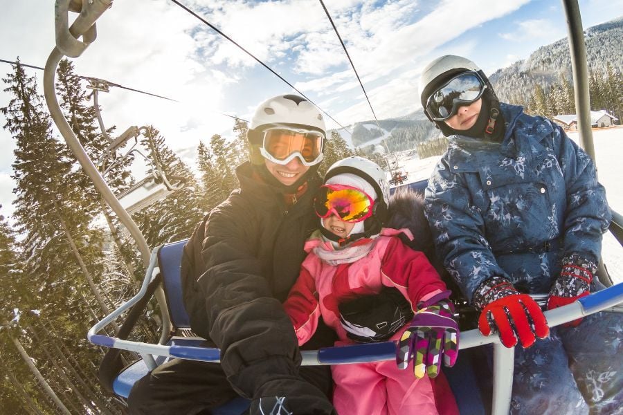 Family on ski lift in googles and snow gear holding selfie stick with mountains and sun behind them. 