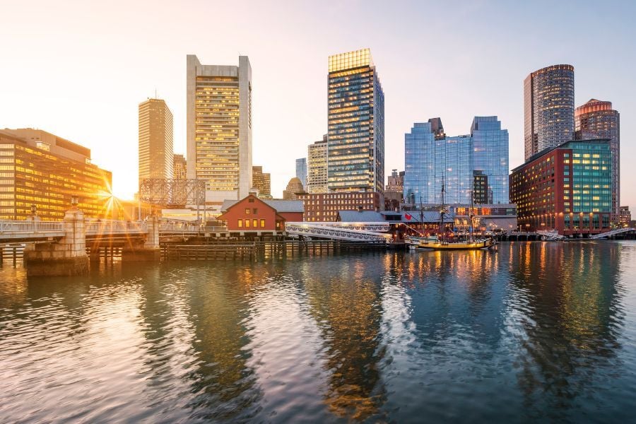 View of Boston harbor with boats in the water and buildings in the background with sun shining through. 