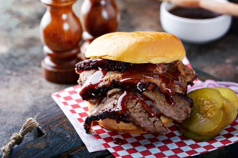 Brisket sandwich with sauce running down next to sliced picked on a sheet of red and white checked paper.