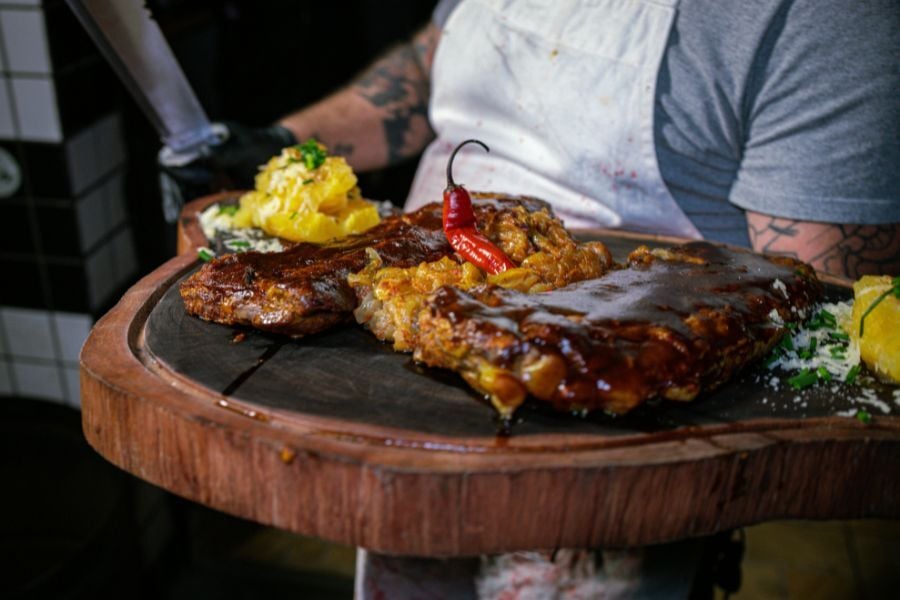 Person with tattooed arms wearing a gray t-shirt and white apron carries block of wood with rack of ribs, peppers, and yellow side on top. 