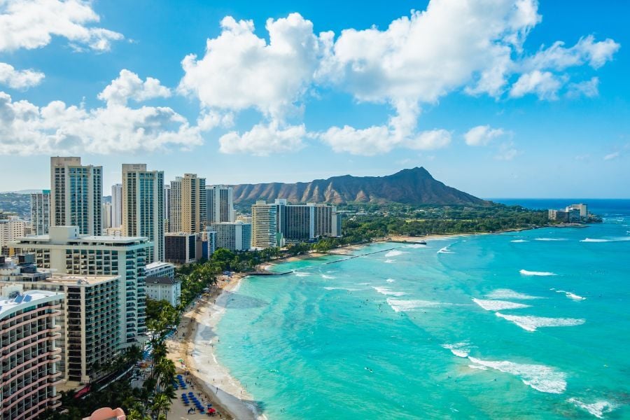 View of Waikiki beach in Oahu, Hawaii with surfers in the water, buildings on the coastline, and Diamond Head in the background. 