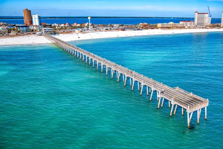 Aerial view of Pensacola Beach pier in vibrant water with beach and buildings behind. 