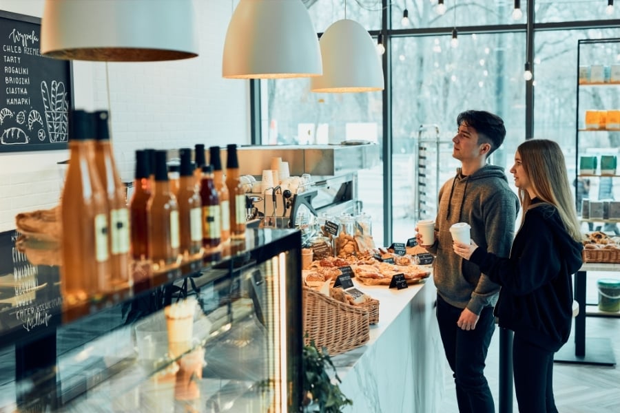 Man and woman in dark neutral clothing stand with to-go coffee cups in hand at coffee shop/bakery counter filled with pastries. 