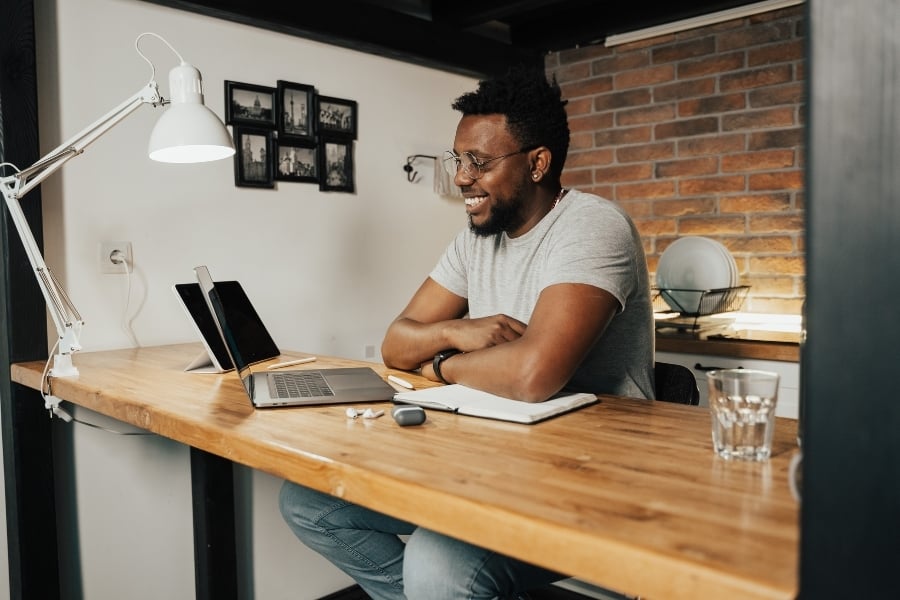Man wearing glasses and gray shirt sits modern room with brick wall and black accents, sits at wooden desk with his arms crossed as he smiles at laptop sitting on table. 