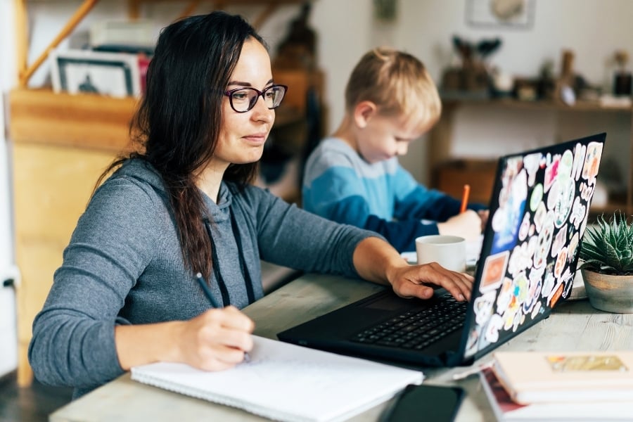 Mom wearing glasses and gray hoodie sits at kitchen table next to to young boy as she takes notes on paper and types of black laptop covered in stickers. 