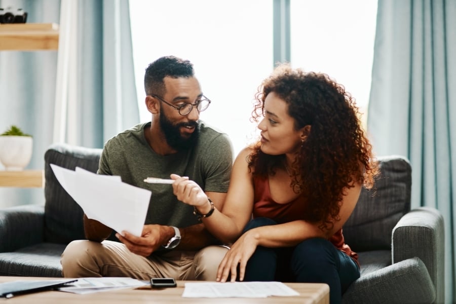 Man in glasses holding papers sits next to woman on couch pointing at papers with pen as they in front of coffee table with more papers and phone/calculator. 