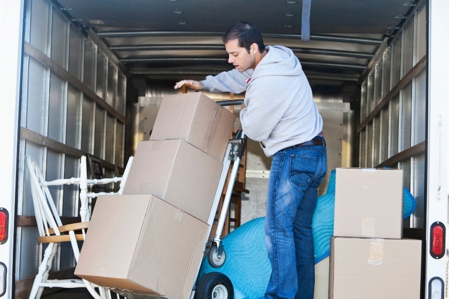 Man in gray hoodie loading cardboard boxes onto truck with dolly.