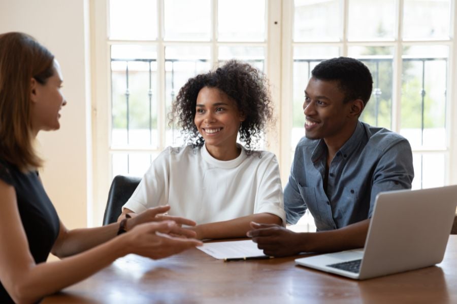 Man in blue button down sits smiling next to woman in white shirt talking to woman across the table gesturing to the laptop on the table..