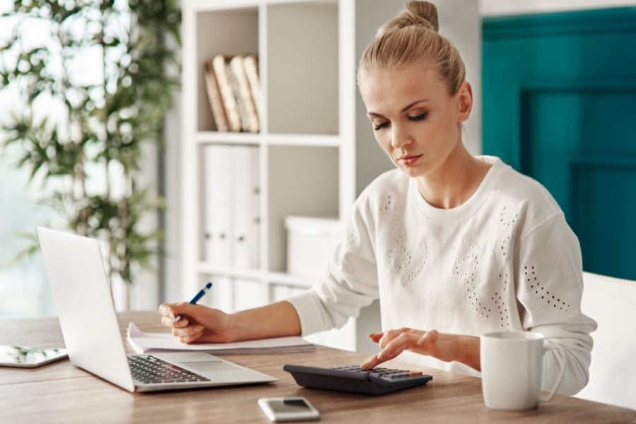 Woman with hair in a bun wearing a long-sleeve white shirt looks at calculator on desk while holding pen. Has a notebook, coffee mug, and computer on desk and plant in background.