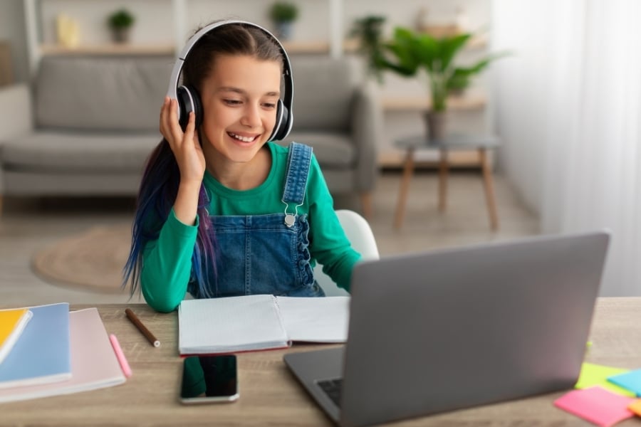 Young girl in green shirt and denim overalls has hand on headphones and smiles at computer on table with phone and notebook by her. 