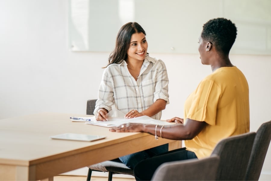 Interview between two women sitting at table smiling at one another.