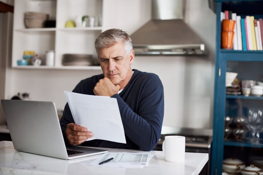 man looking at paperwork while sitting at table with laptop and coffee cup