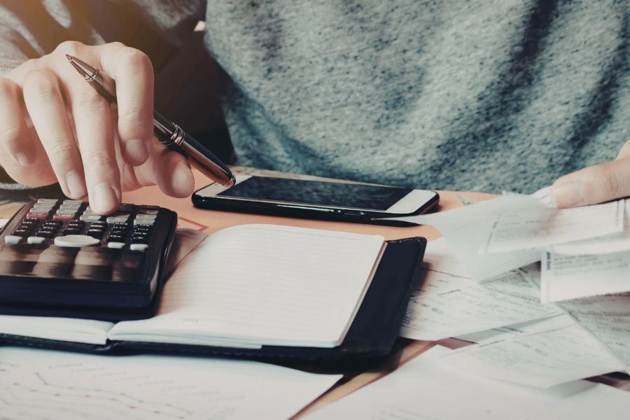Person in long sleeve shirt sits at table with receipts, calculator, notebook, pen, and phone in front of them. 