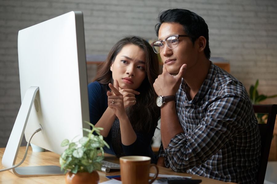 Man in glasses wearing watch and woman sitting at desk looking closely at computer in front of them. 
