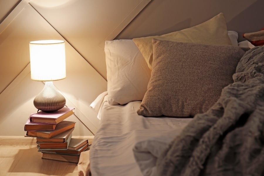 Cozy, neutral bed on light-colored wooden floor with stack of books topped with a small lamp sitting nearby. 
