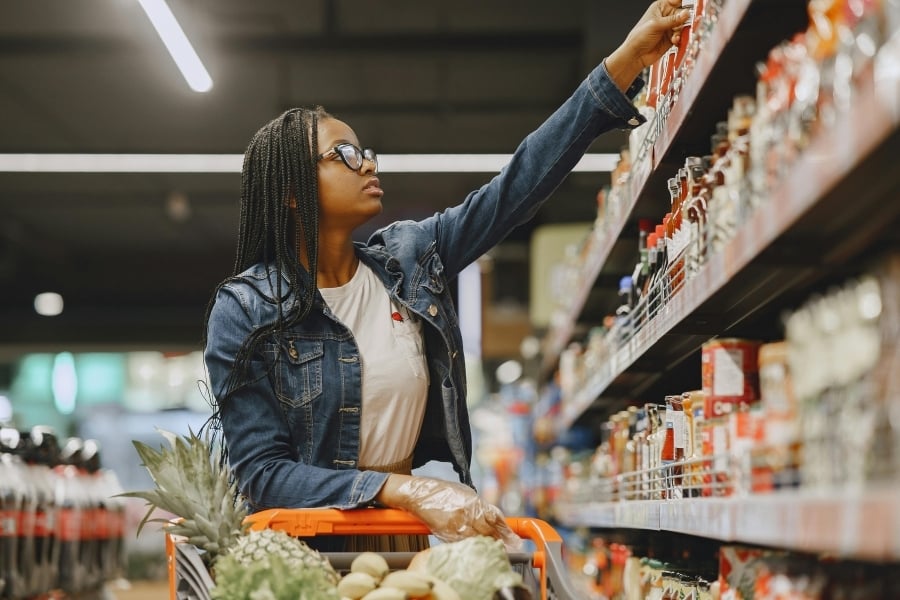 Woman wearing denim jacket, glasses, and gloves pushes orange shopping cart and reaches for item on top shelf in grocery store. 