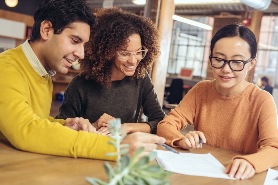 Landlord in orange sweater and glasses meet with male in yellow sweater and female in glasses at table with paperwork and pen in hand.