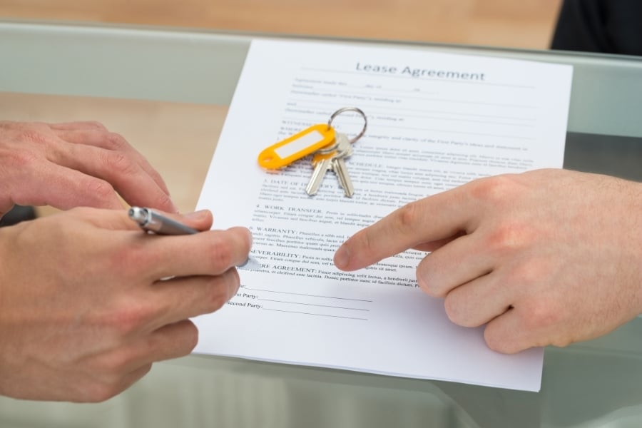 Person holding pen to sign lease agreement on desk with yellow keys sitting on top with other person pointing to the line for them to sign. 