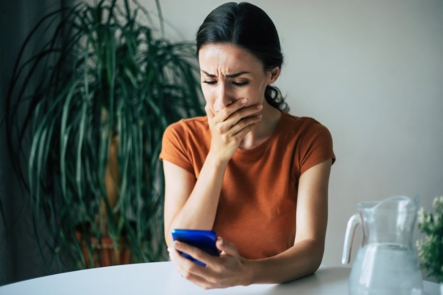 Woman in orange shirt holds hand over her mouth looking distressed as she looks at the blue phone she's holding. She's sitting at white table with pitch of water next to her and green palm tree behind her. 