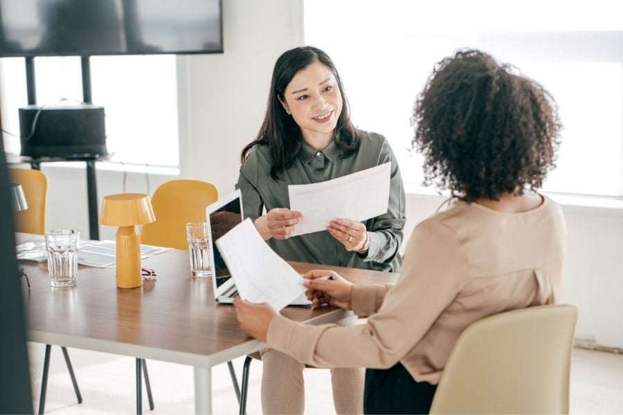 Two women in business attire sitting across table from one another talking about papers.