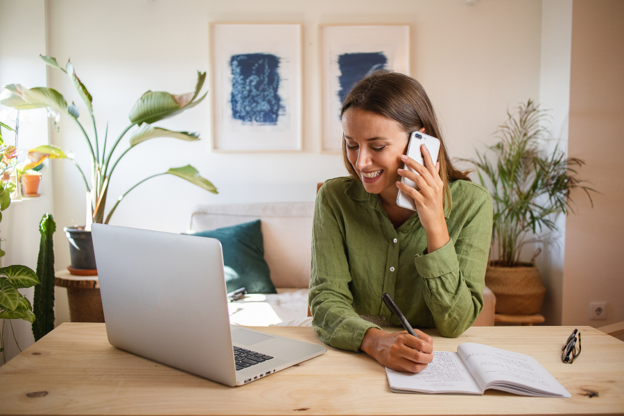 Woman in green long sleeve button down takes notes in notebook while on the phone and looking at computer.