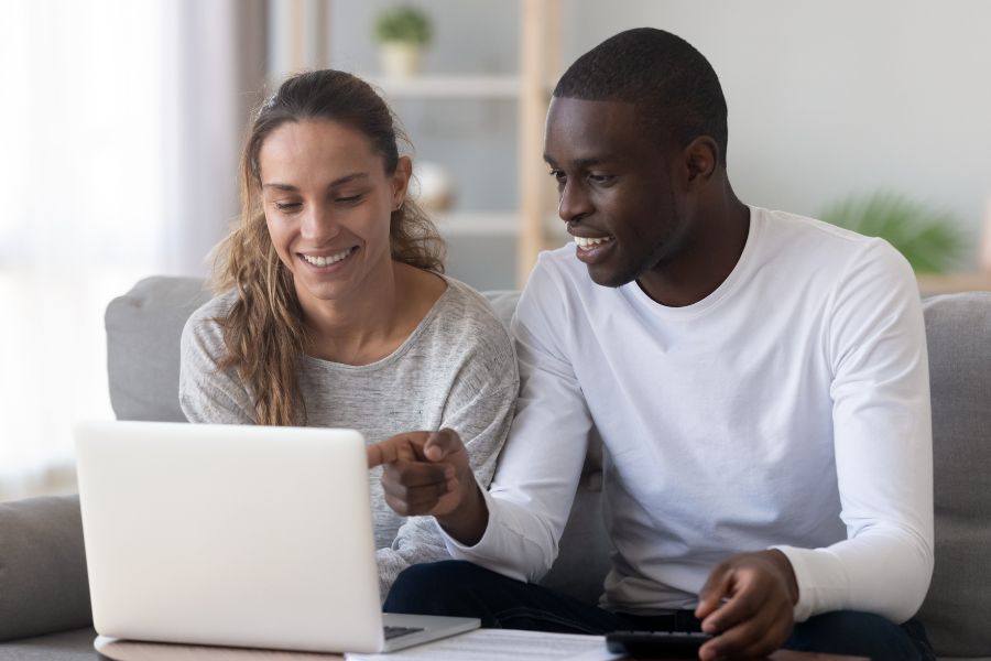Man and woman in neutral long sleeve shirts sit pointing and smiling at computer in front of them. 