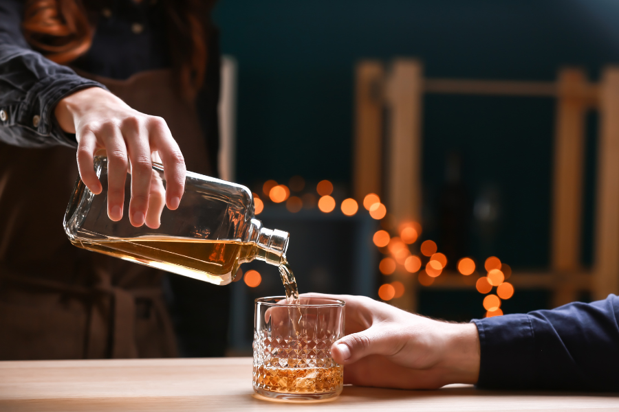 Bartender pouring whiskey into patron's glass with string lights blurred in the background. 