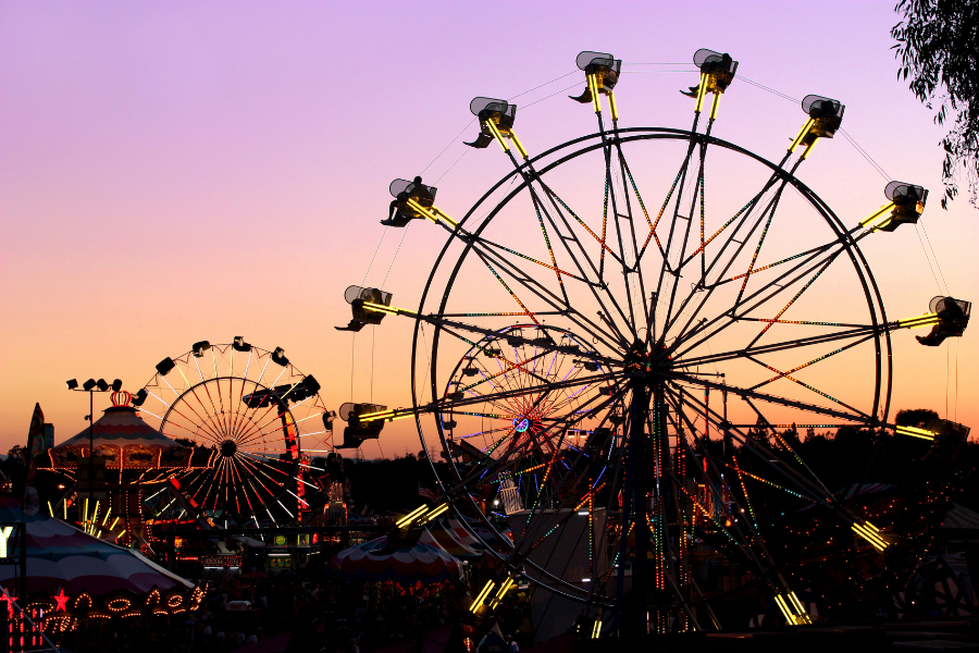 Night at fall state fair with lights on ferris wheels shining as the sun goes down casting an orange/purple glow in the sky. 