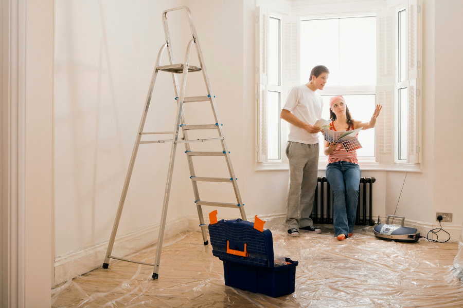Couple perched near radiator in house with plastic-covered floors, tools nearby, and ladder set up to start home repairs. 