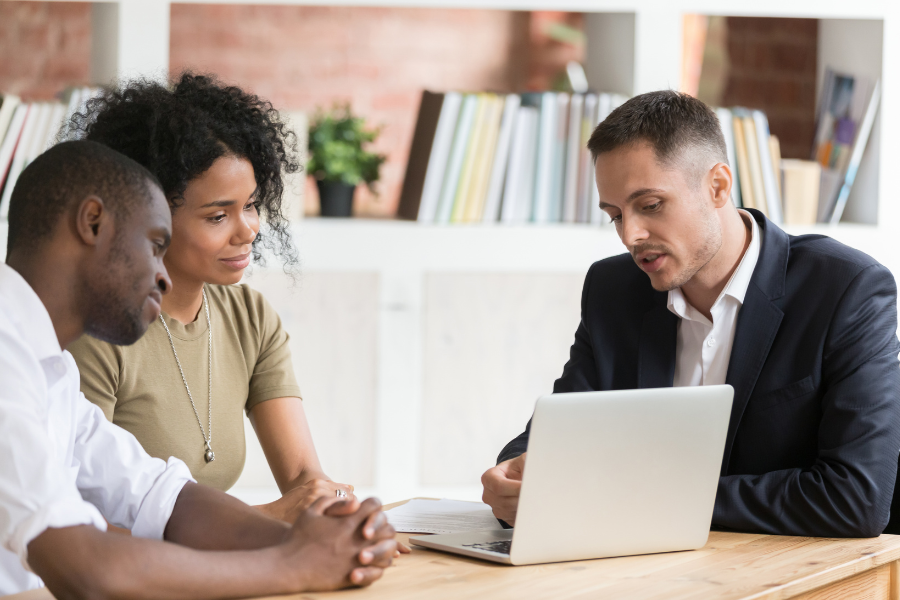 Couple sit forward at table with closing agent in suit pointing to laptop in front of them.