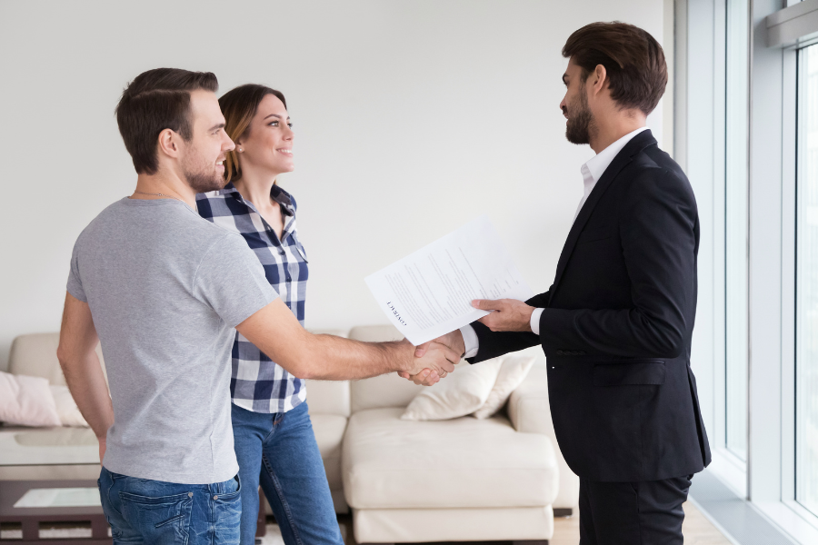 Real estate agent in suit shakes hands with hand and woman in living room with beige couch.