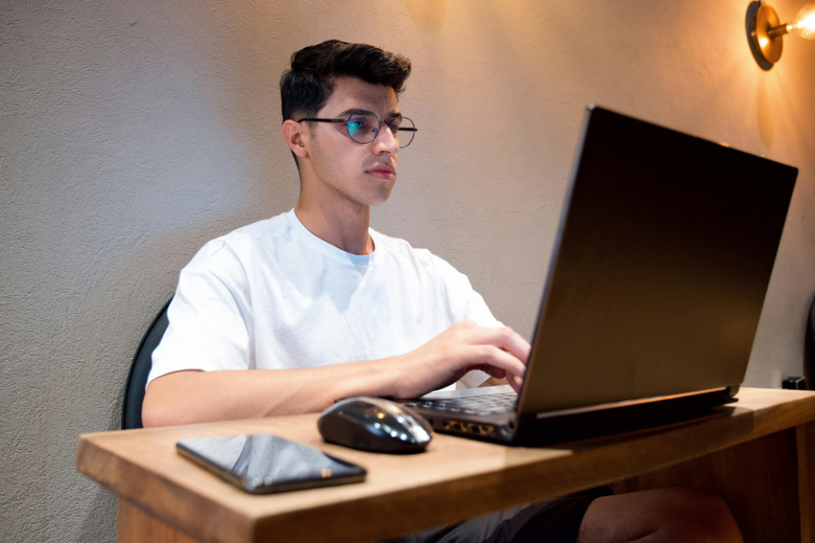 Man wearing glasses and white t-shirt sits at wooden desk and types on computer