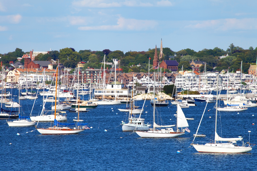 Sail boats in harbor in Newport, RI with brick buildings and trees on shore behind. 