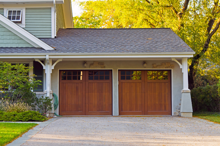 Wooden and glass garage doors attached to sage green home with lit trees behind. 