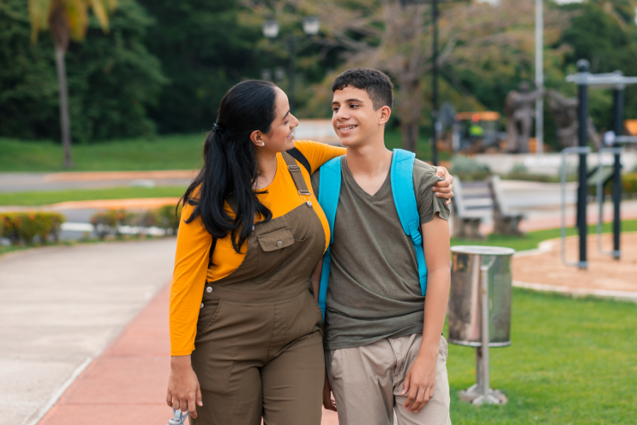 Mom in brown overalls walks with arm around son wearing backpack smiling