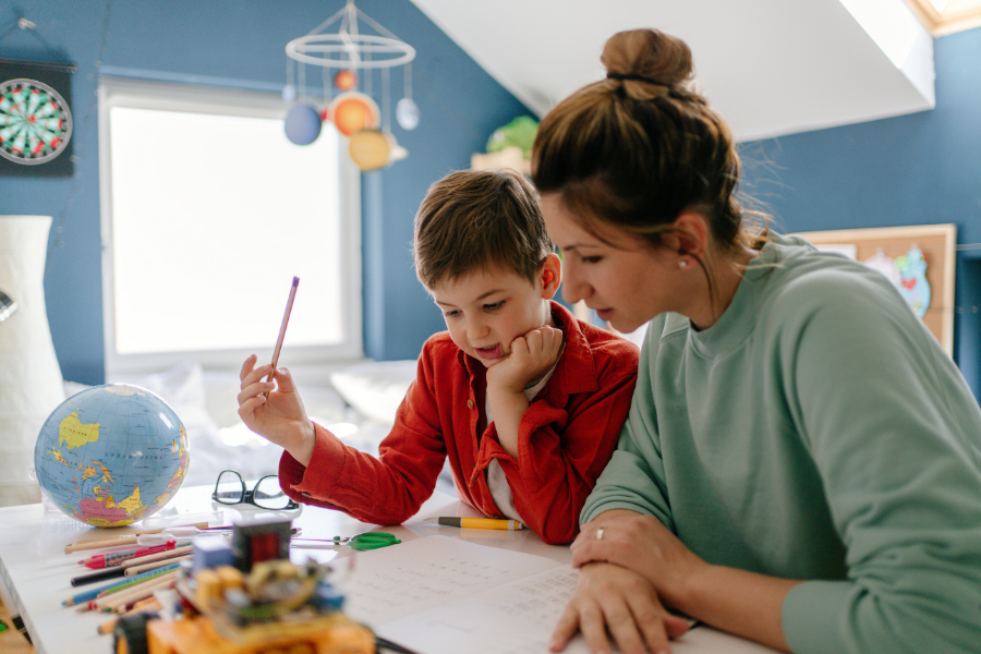 Mom wearing green sweatshirt helps son in red shirt with school work in blue room with globe and solar system. 