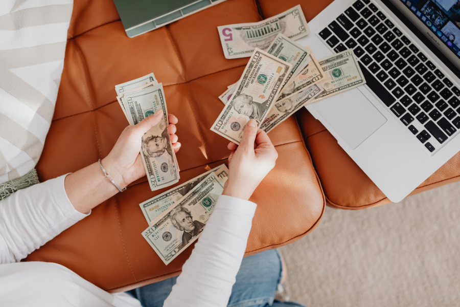 Woman counting cash on orange couch next to laptop. 